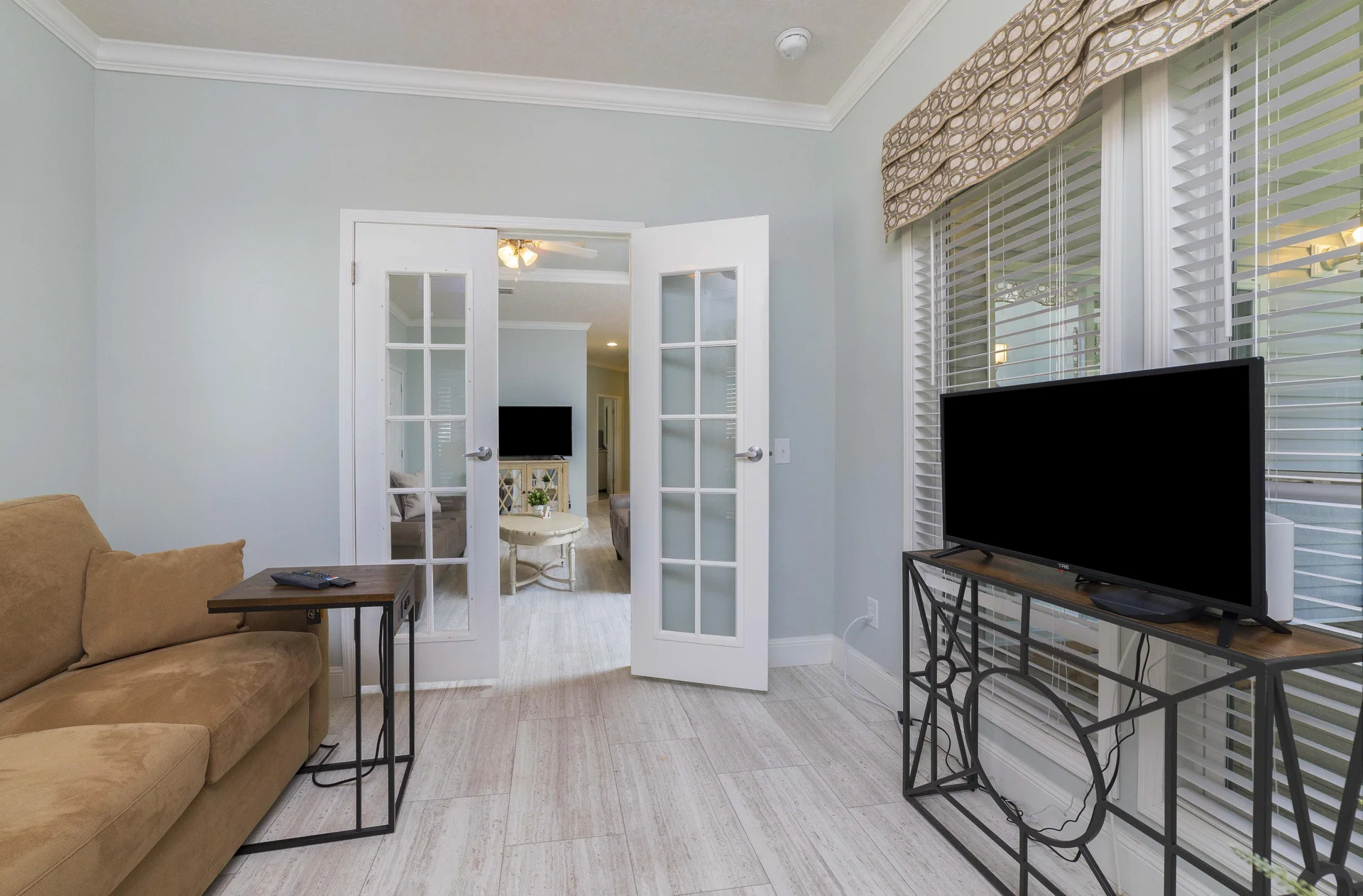 Small sitting area with tan sofa, black metal side table, and TV on decorative metal console by windows.