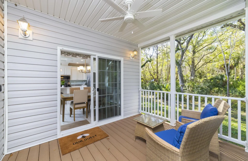Covered porch with wicker chairs featuring blue cushions and a glass-topped side table overlooking trees.