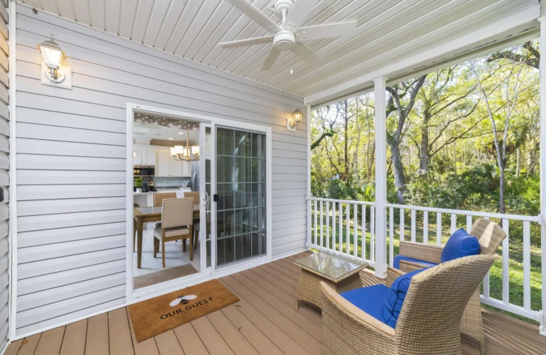 Covered porch with wicker chairs featuring blue cushions and a glass-topped side table overlooking trees.