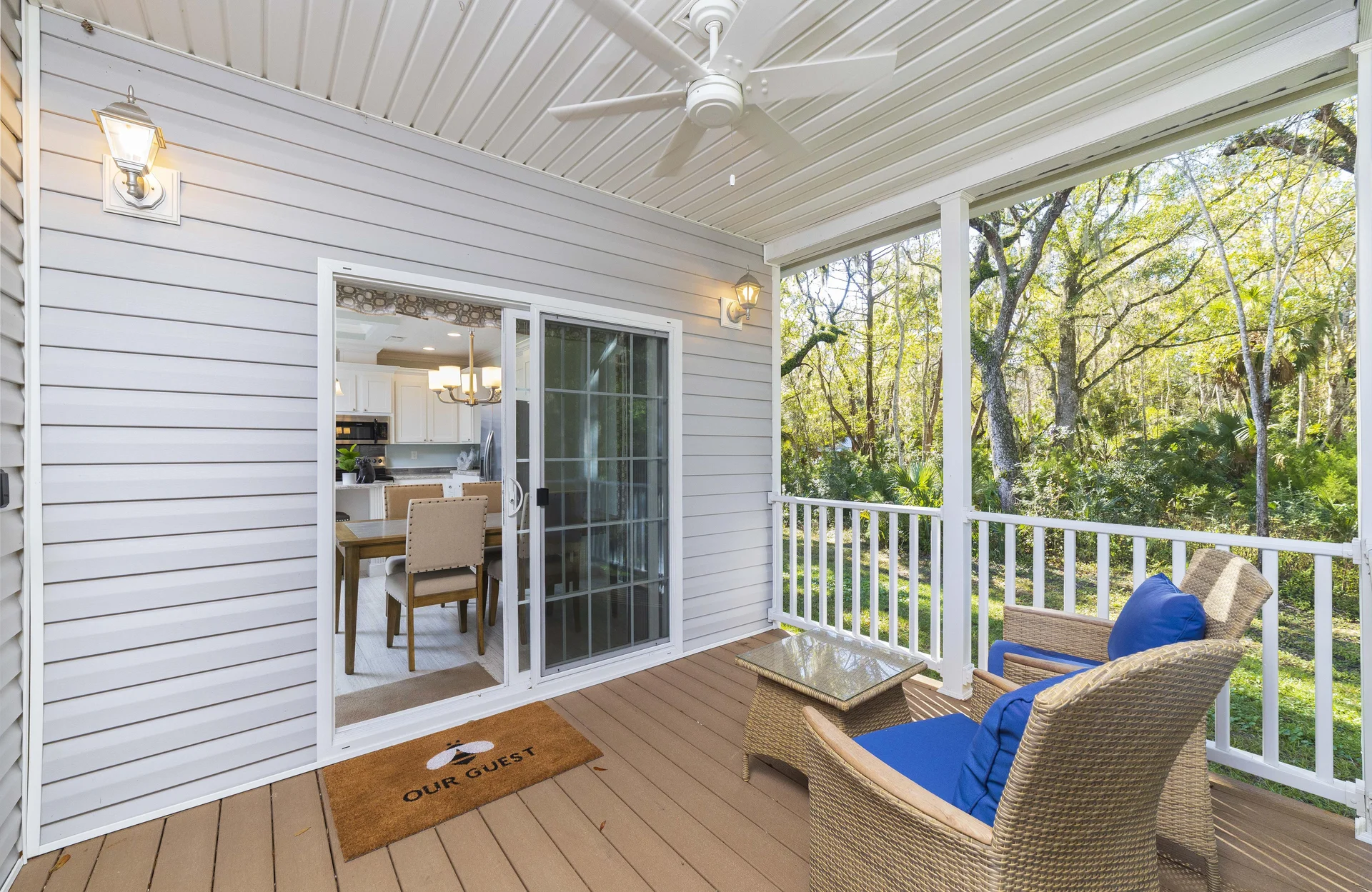 Covered porch with wicker chairs featuring blue cushions and a glass-topped side table overlooking trees.