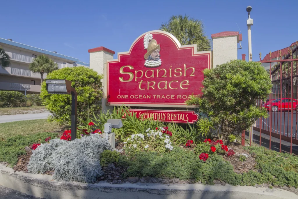 Red and gold Spanish Trace entrance sign stands amid green shrubs and colorful flowers by gated parking.