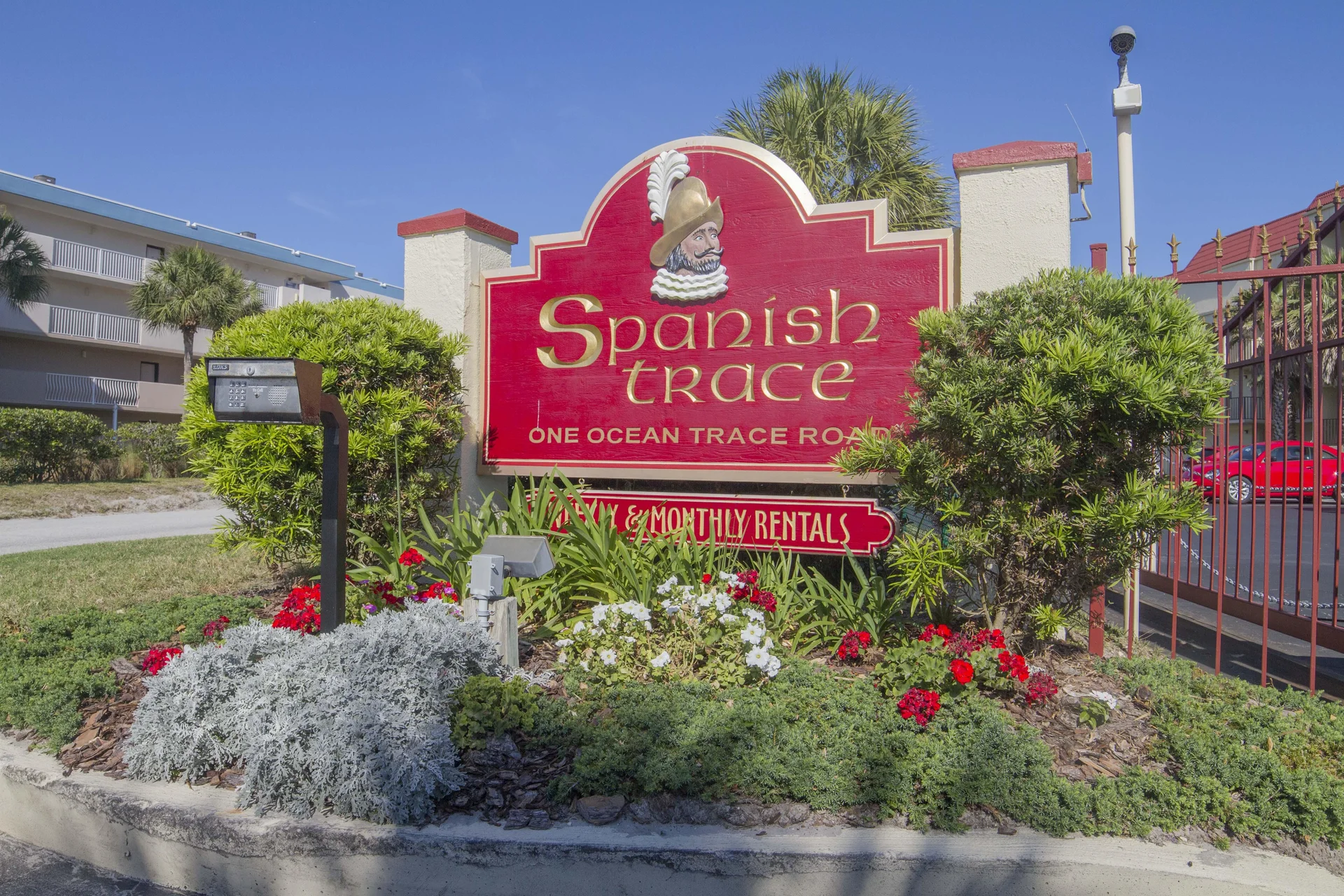 Red and gold Spanish Trace entrance sign stands amid green shrubs and colorful flowers by gated parking.
