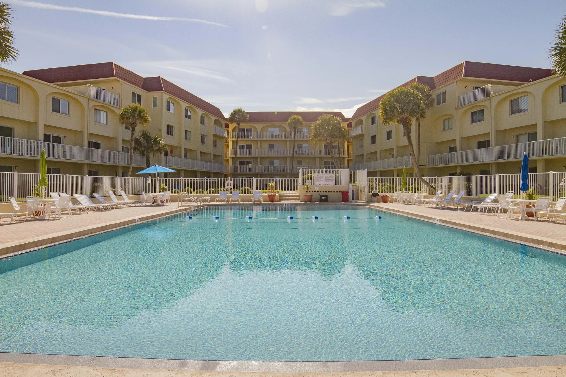 Large rectangular swimming pool surrounded by white lounge chairs, umbrellas, and palm trees against a beige condo complex.