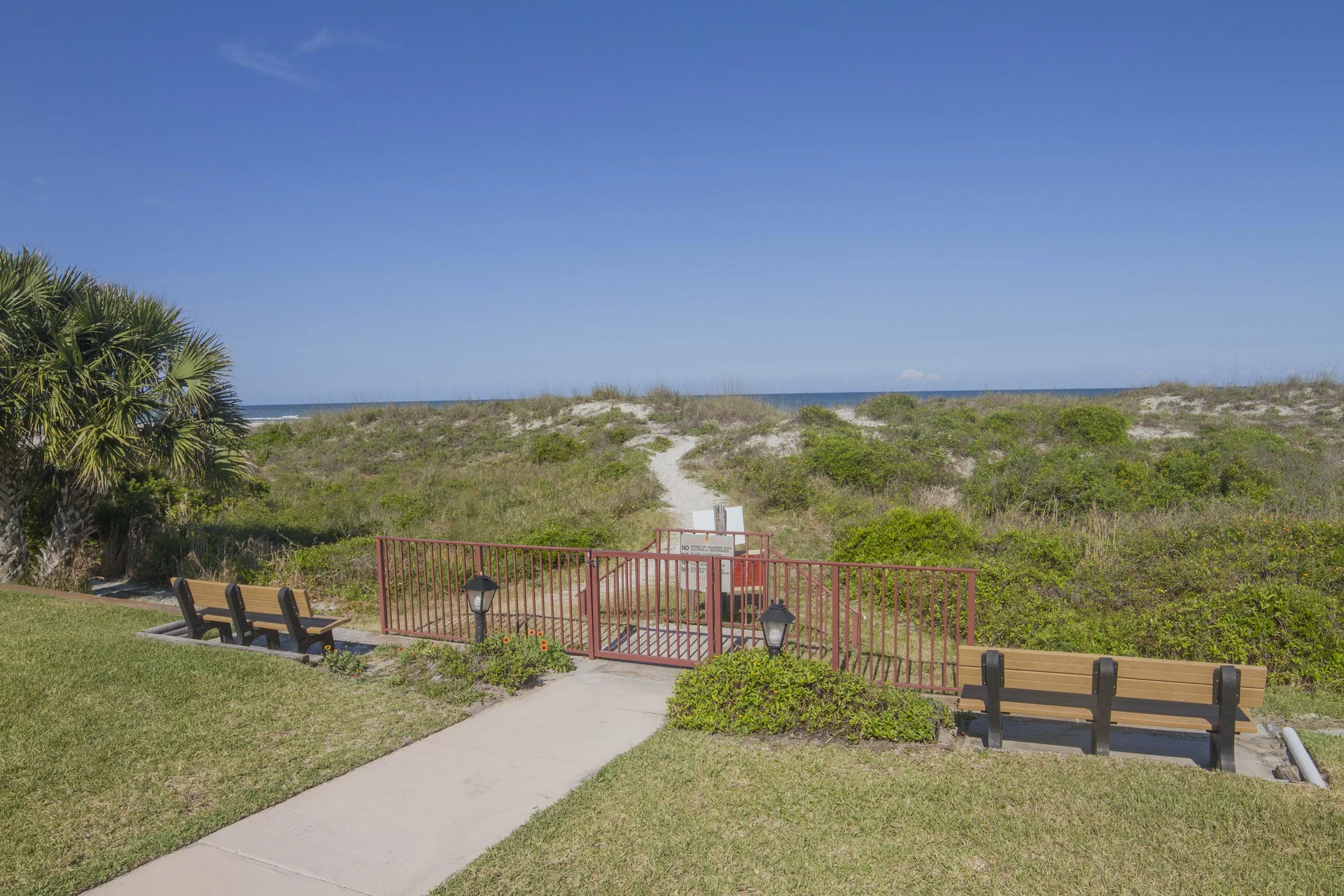 Concrete pathway leads past wooden benches to a red metal gate opening onto a sandy dune trail toward the ocean.