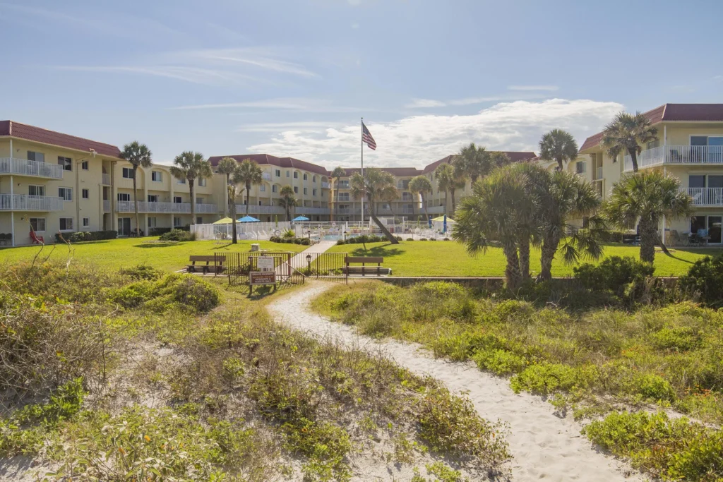 Sandy path leads from coastal vegetation to a grassy courtyard with benches and a pool beyond the condo buildings.