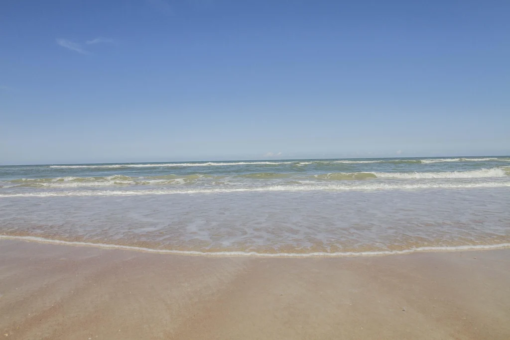 Gentle waves roll onto a smooth sandy beach beneath a clear blue sky at the ocean’s edge.