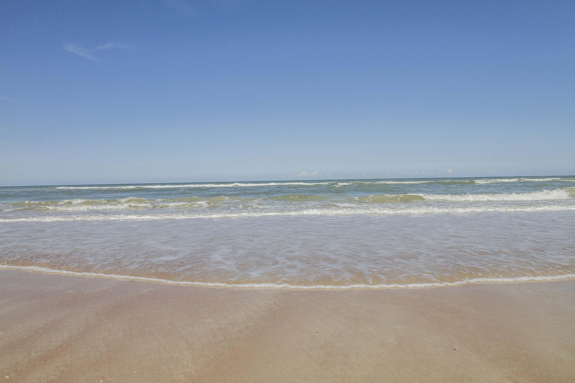 Gentle waves roll onto a smooth sandy beach beneath a clear blue sky at the ocean’s edge.