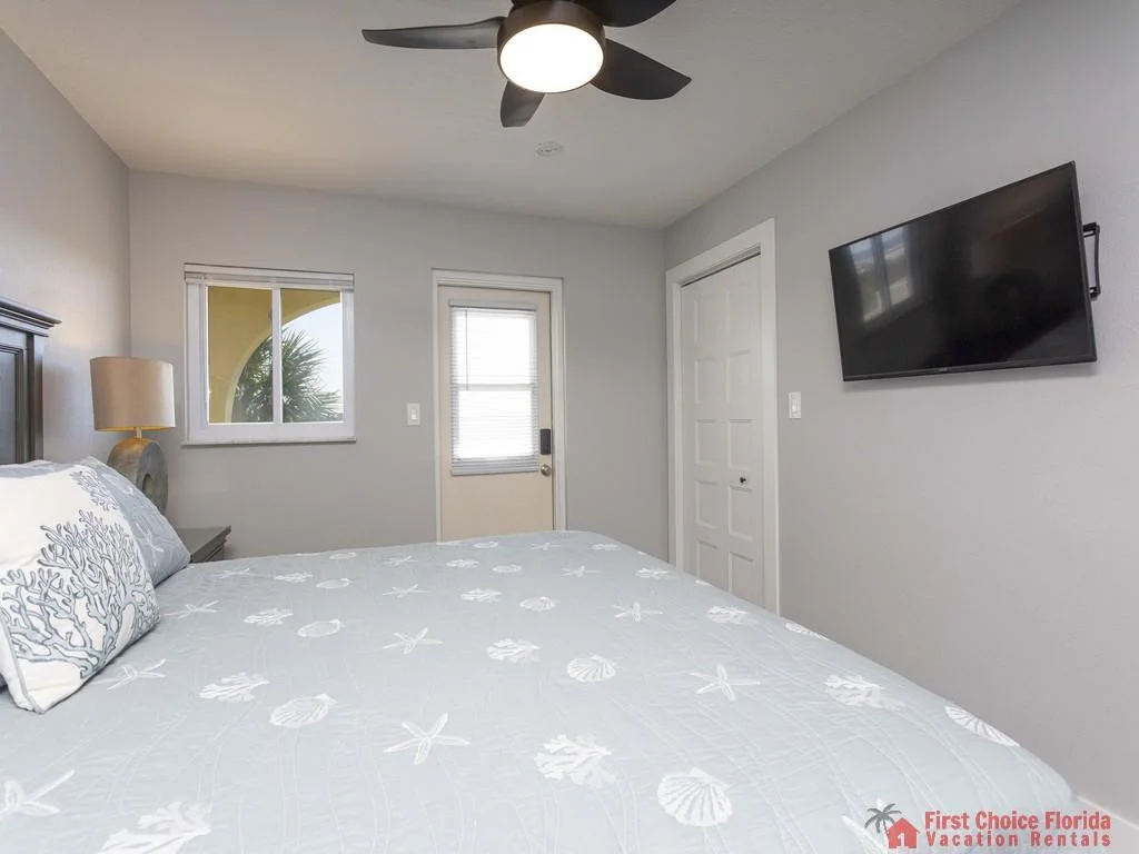 Bedroom with light gray walls, patterned blue bedding, ceiling fan, wall-mounted TV, and door to balcony.