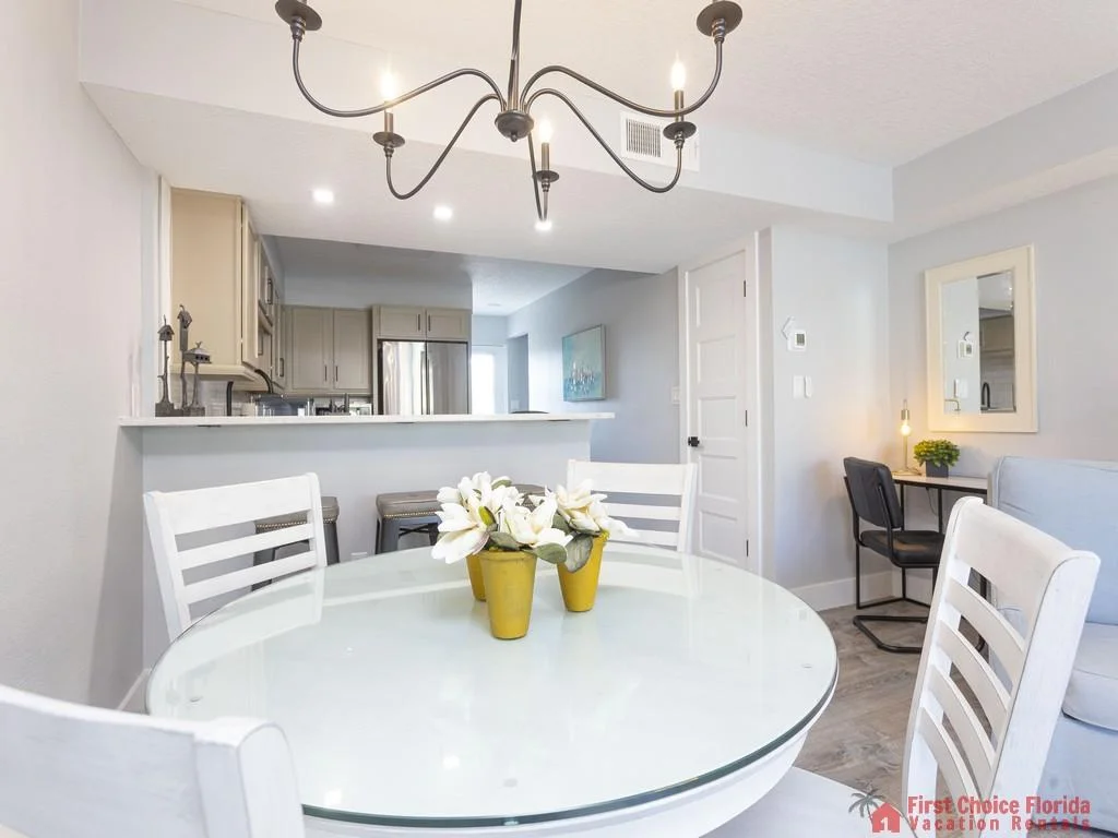 Round glass dining table with white wooden chairs beneath a black metal chandelier, adjacent to a compact workspace.