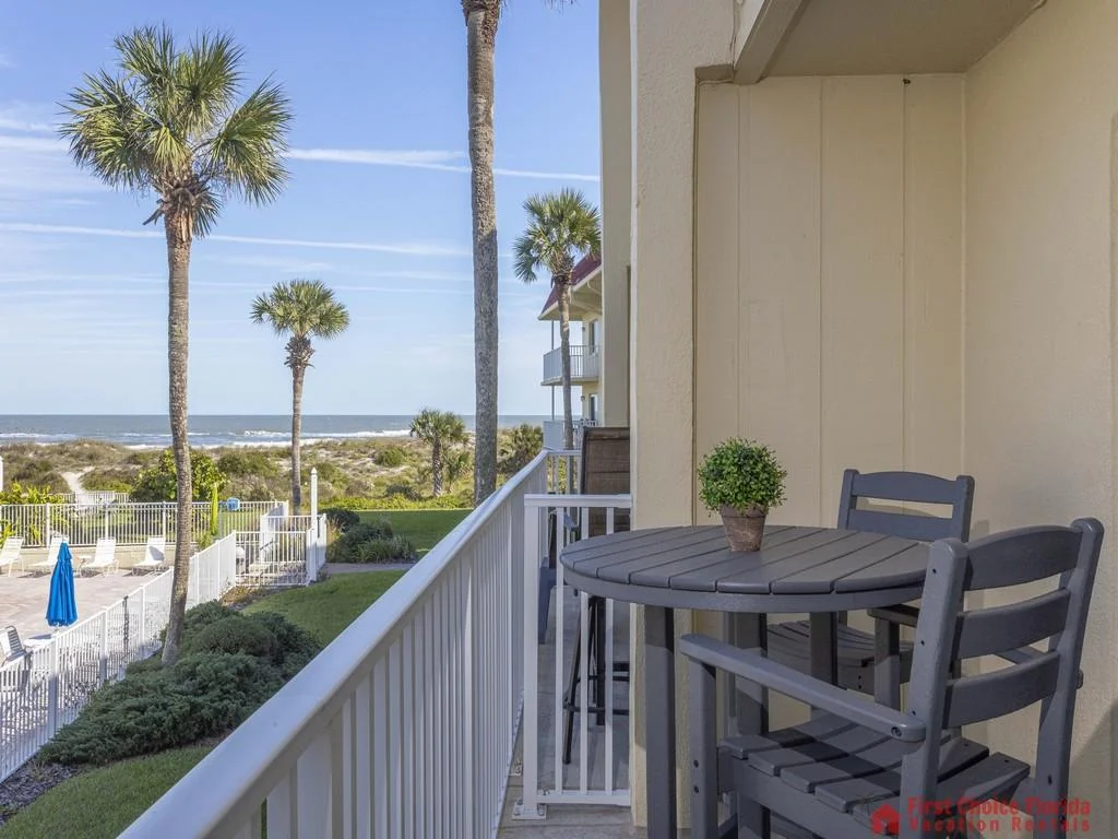 Outdoor balcony features a round gray table with matching chairs and a small potted plant, overlooking palm trees and ocean.