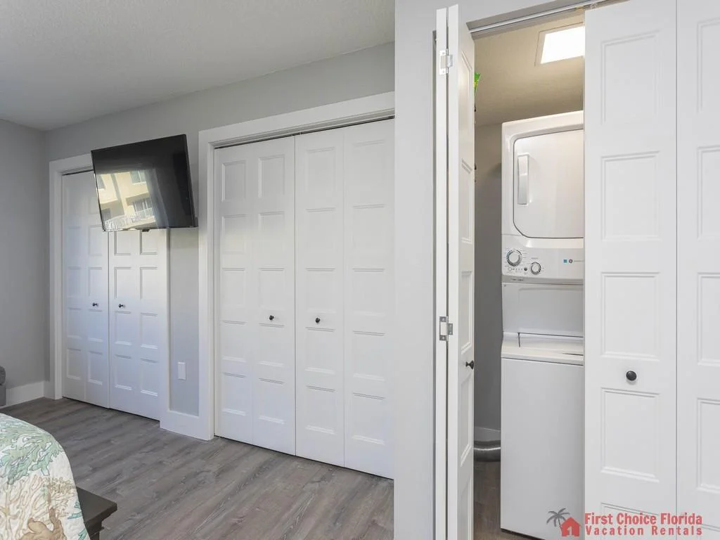 Laundry closet with stacked washer and dryer beside white paneled bedroom closets and wall-mounted TV.