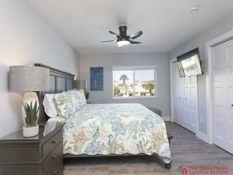 King bed with coral-patterned quilt faces a wall-mounted TV and dual white closet doors in the bedroom.