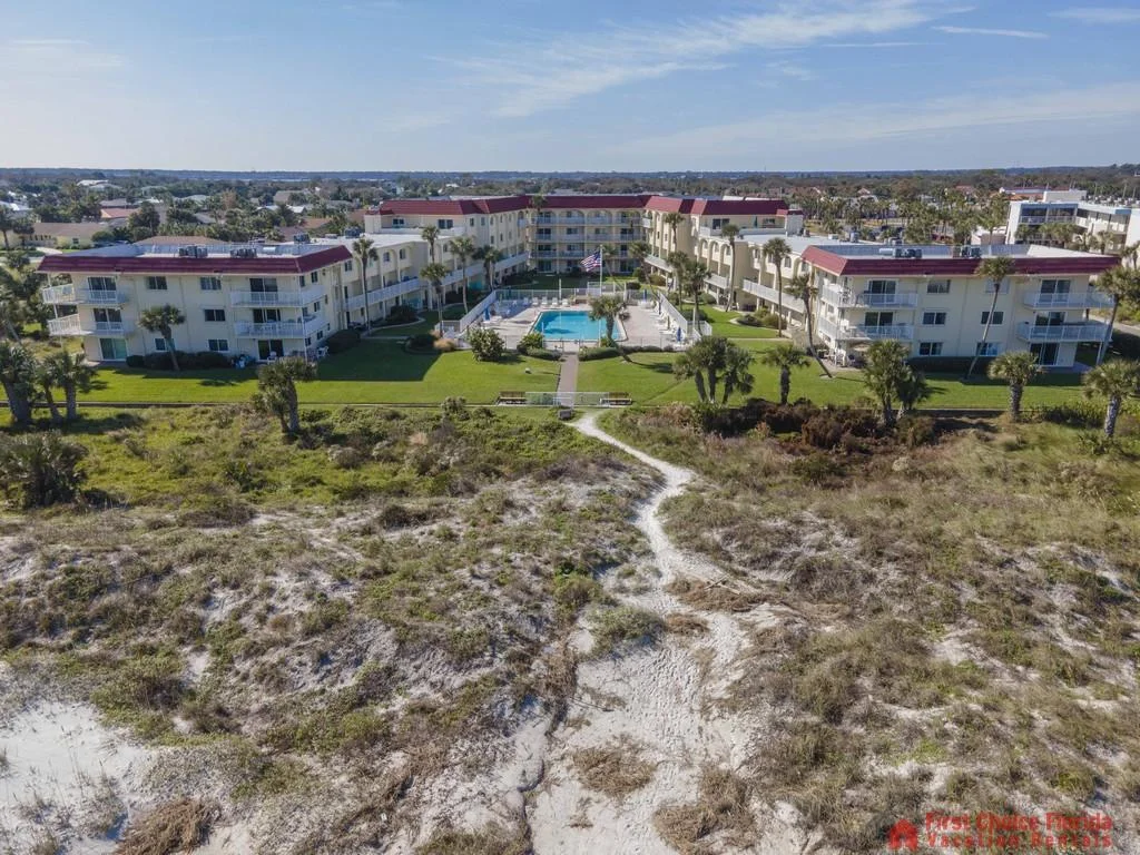 Aerial view of three-story condo buildings with red roofs surrounding a fenced pool, set behind sandy dunes.