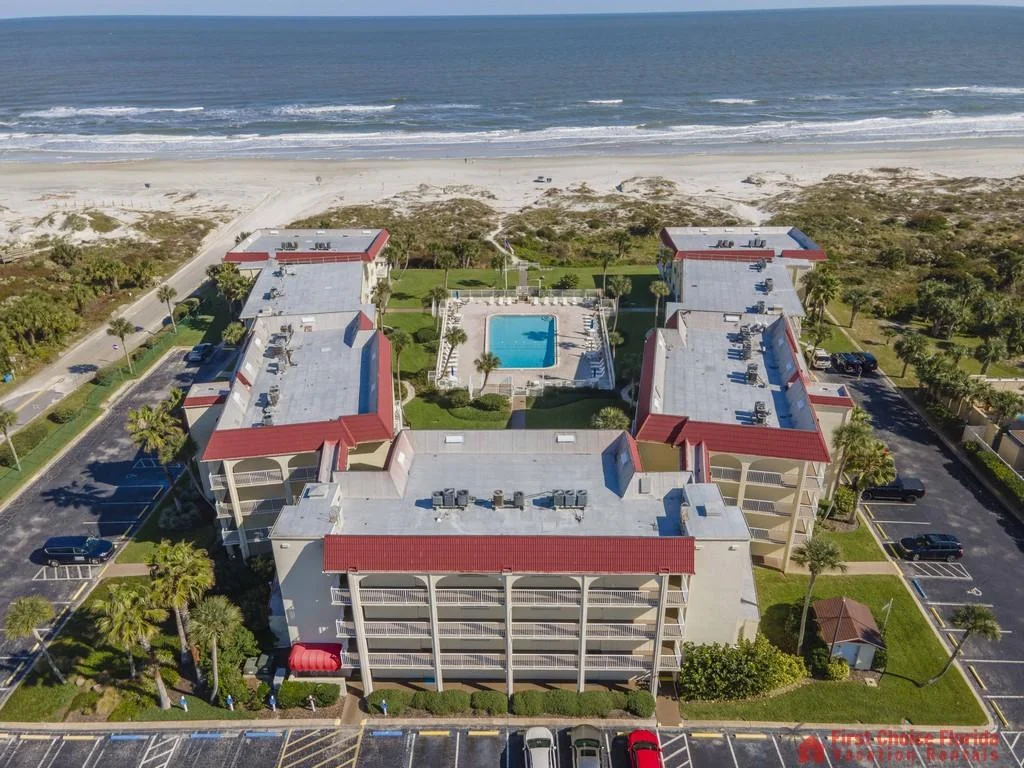 Aerial view of a U-shaped condo complex with red trim, central pool, palm trees, and sandy beach beyond.