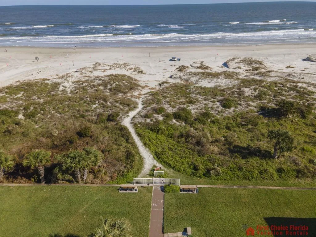 Grassy lawn leads to a sandy path cutting through dunes towards the wide beach and ocean waves.