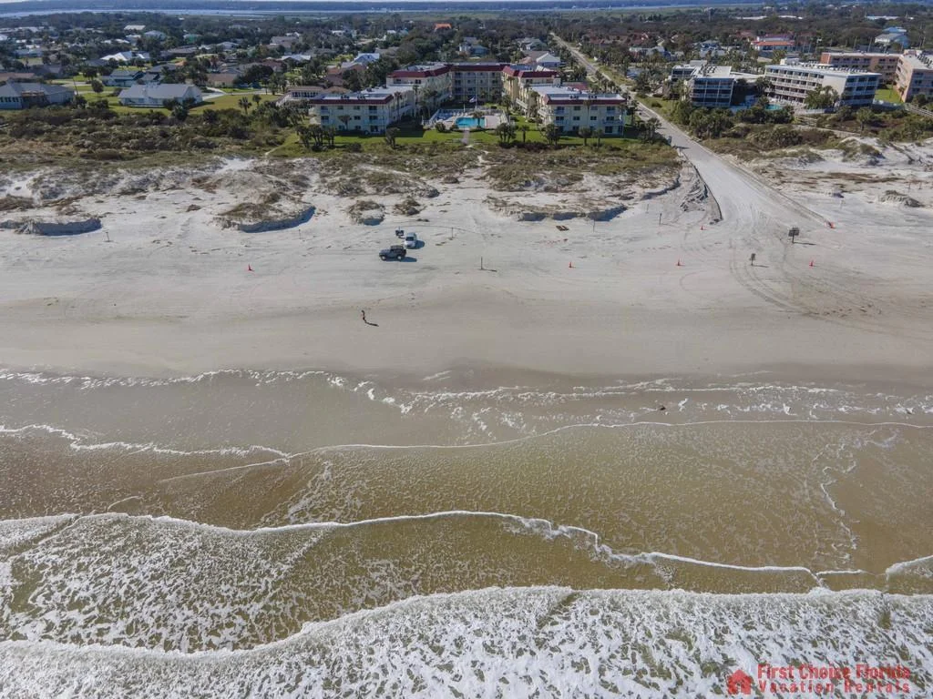 Aerial view of sandy beach with gentle waves, beach access road, and low-rise condos beyond the dunes.