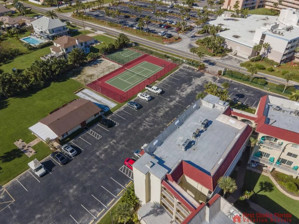 Aerial view of tennis courts, parking lot, and nearby buildings with green lawns and palm trees.
