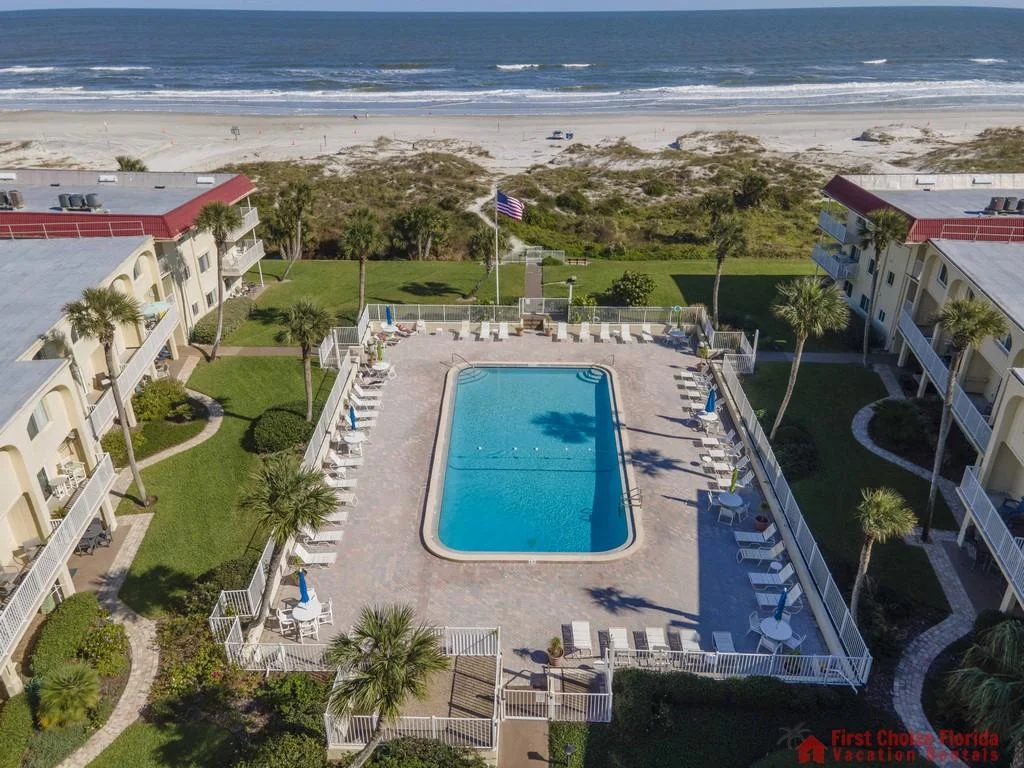 Rectangular pool with white lounge chairs and blue umbrellas sits between two beige three-story condo buildings.