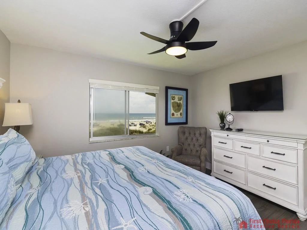Bedroom with coastal-themed bedding, tufted armchair, white dresser, wall-mounted TV, and ocean view through window.