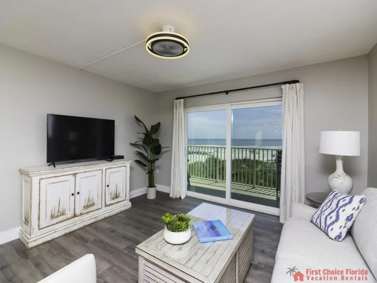 Flat-screen TV atop distressed white cabinet faces a light gray sofa with patterned pillows near sliding glass doors.
