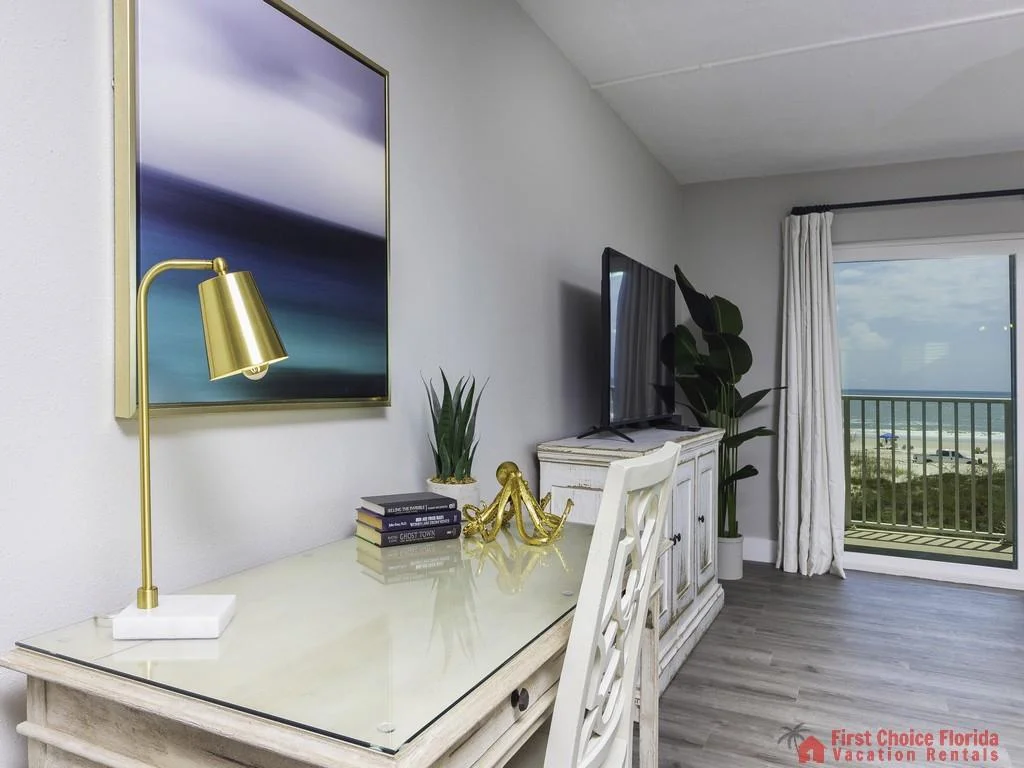 Glass-top desk with brass lamp and stacked books faces a distressed white media cabinet holding a flat-screen TV.