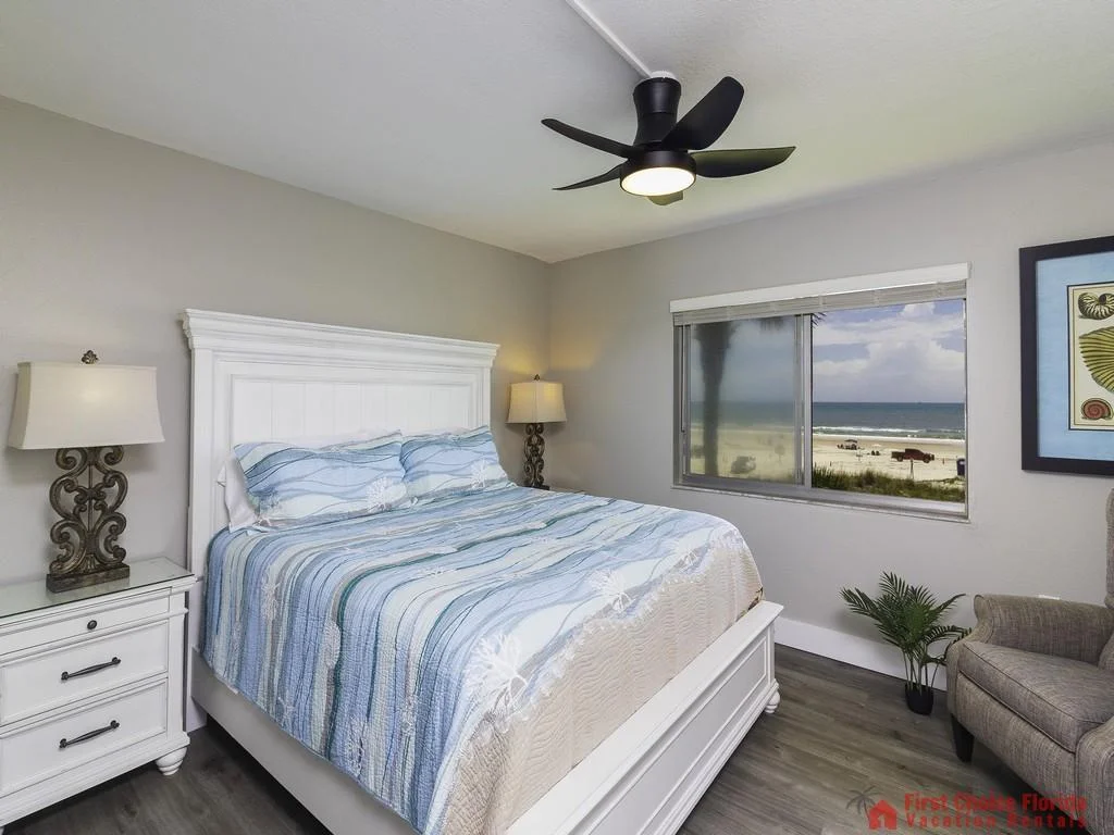 White wooden bed with blue-striped bedding faces a window showcasing a sandy beach and ocean waves.