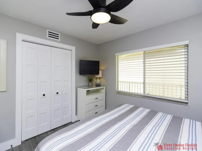 Striped gray and blue bedding covers a white bed beneath a ceiling fan in the bedroom with a window and dresser.