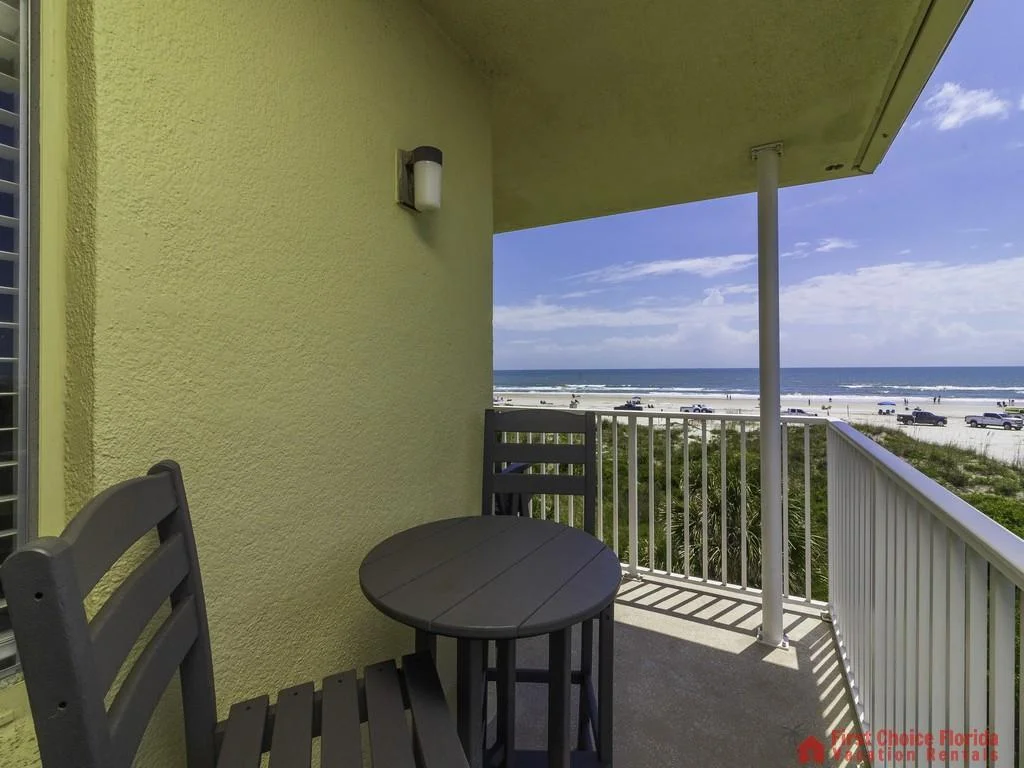 Textured yellow exterior walls frame a small balcony with dark wooden chairs and round table overlooking the beach.