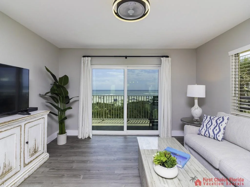Sliding glass doors open onto a balcony with ocean views beyond a light gray sofa and rustic white TV cabinet.