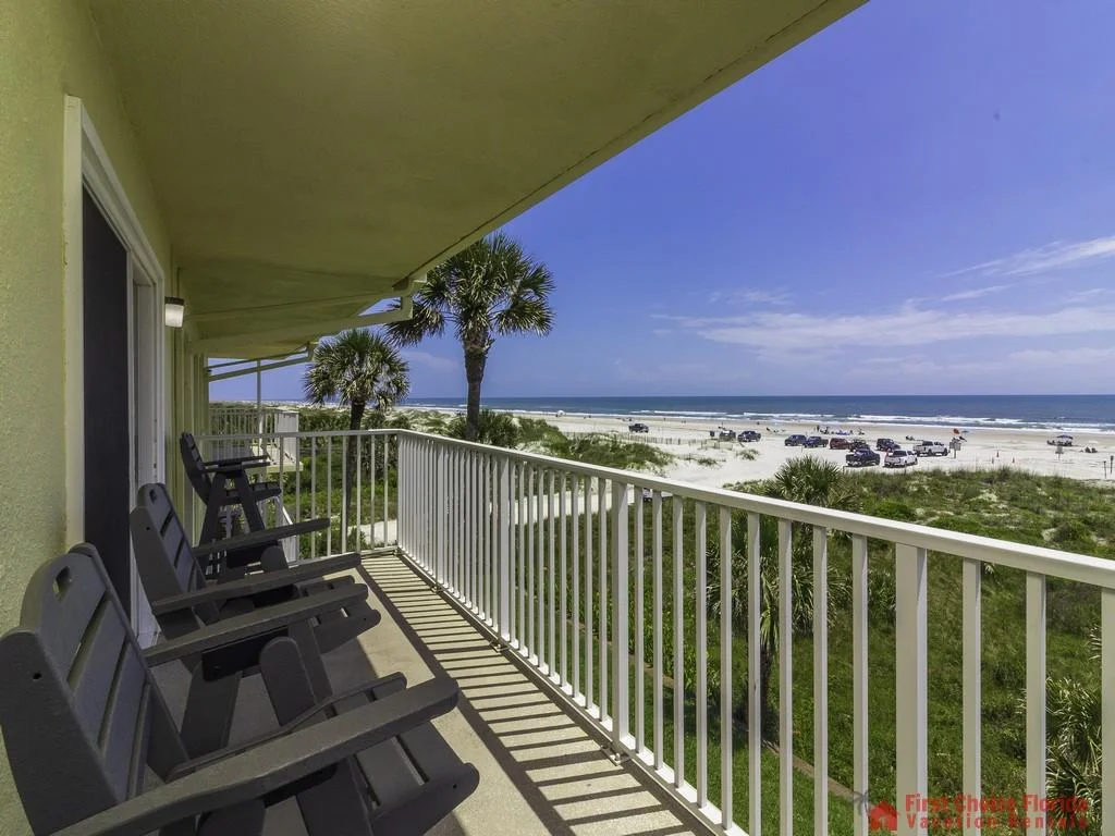 Balcony lined with dark Adirondack chairs overlooks palm trees, sandy beach, and ocean waves under clear sky.