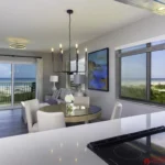 Glass-top dining table surrounded by beige chairs sits beside a window revealing ocean views.