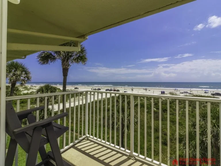 Concrete balcony floor shaded by overhead structure, looking past palm trees toward sandy beach and blue ocean.