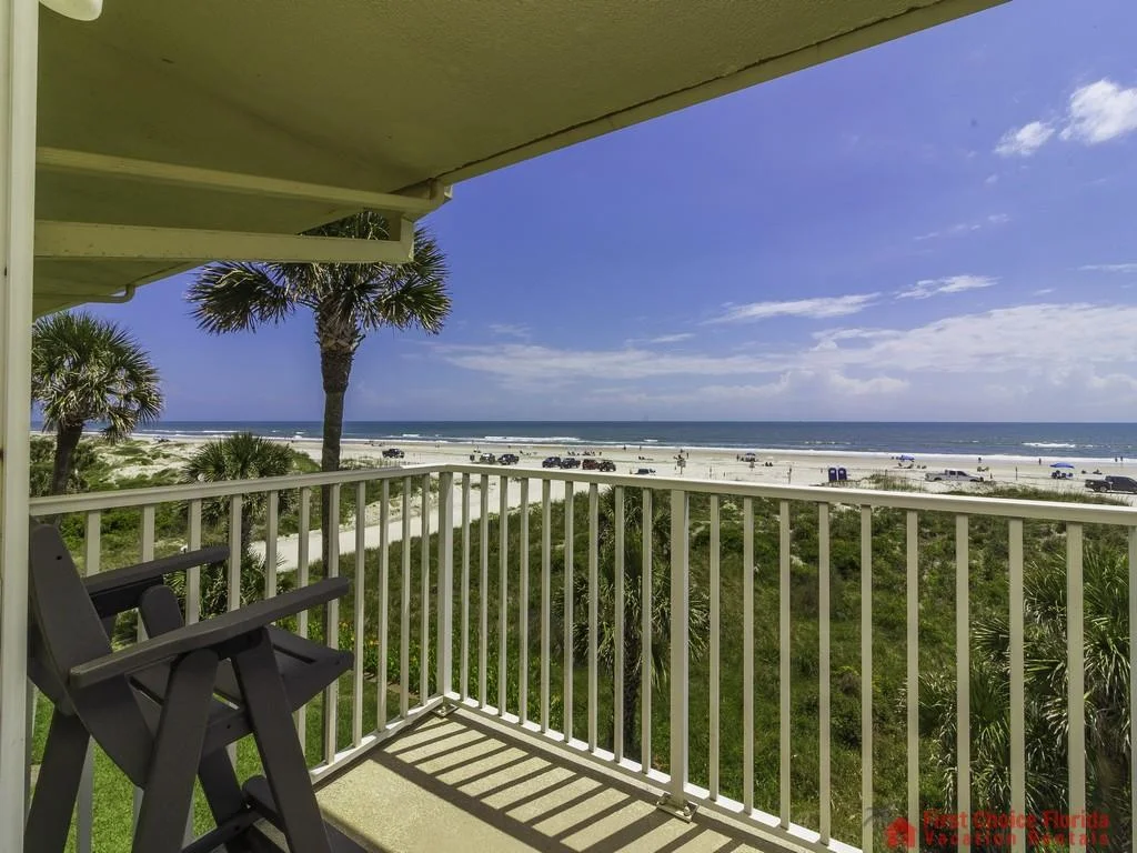 Concrete balcony floor shaded by overhead structure, looking past palm trees toward sandy beach and blue ocean.