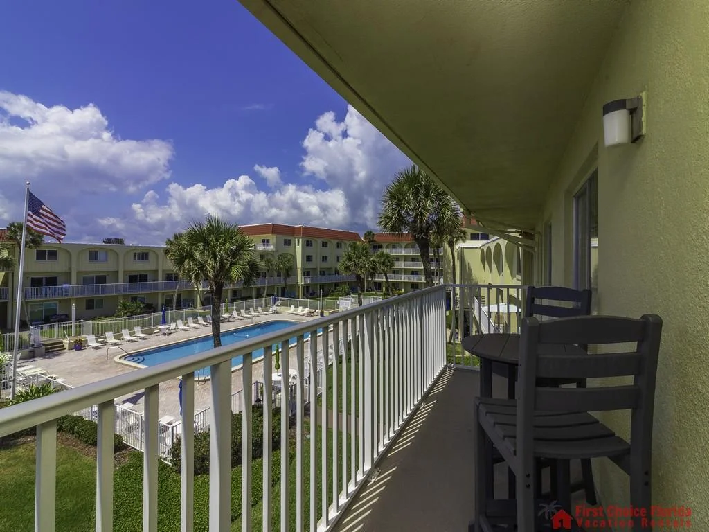 Balcony with dark wooden chairs and table overlooks pool area, palm trees, and neighboring buildings under blue sky.