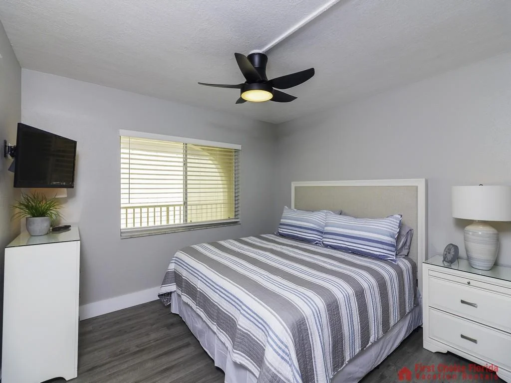 Bedroom with striped bedding, white framed headboard, gray wood floors, ceiling fan, and wall-mounted TV.