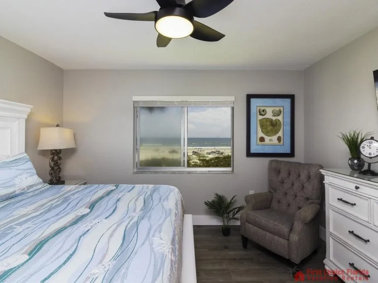 Tufted gray armchair sits beside a white dresser under a wall-mounted TV in the bedroom with beach view window.