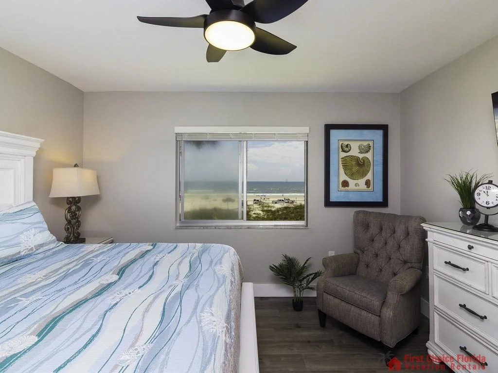 Tufted gray armchair sits beside a white dresser under a wall-mounted TV in the bedroom with beach view window.