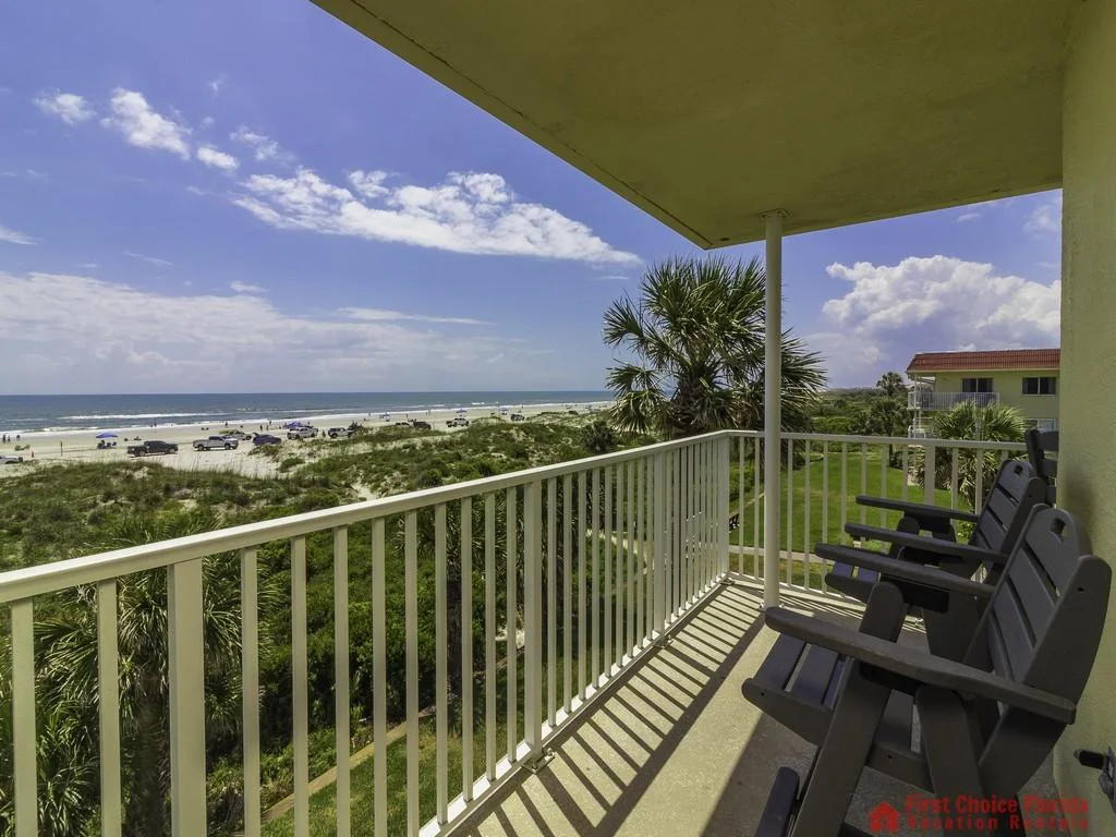 Concrete balcony floor with black Adirondack chairs faces sand dunes, palm trees, and beach parking under a partly cloudy sky.