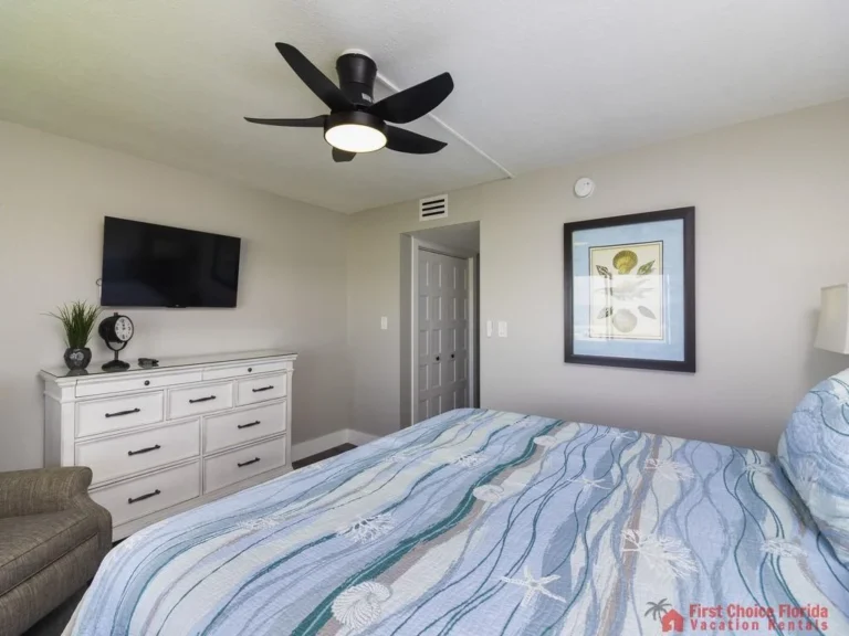 Blue and white seashell-patterned bedding covers the bed beneath a black ceiling fan and wall-mounted TV.
