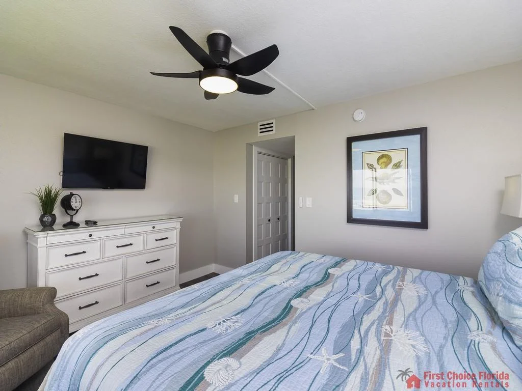 Blue and white seashell-patterned bedding covers the bed beneath a black ceiling fan and wall-mounted TV.
