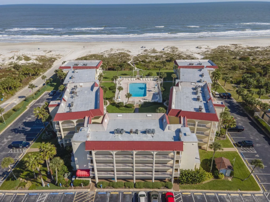 Aerial view of beachfront condo complex with red-tiled roofs, central pool, palm trees, and sandy ocean shoreline.