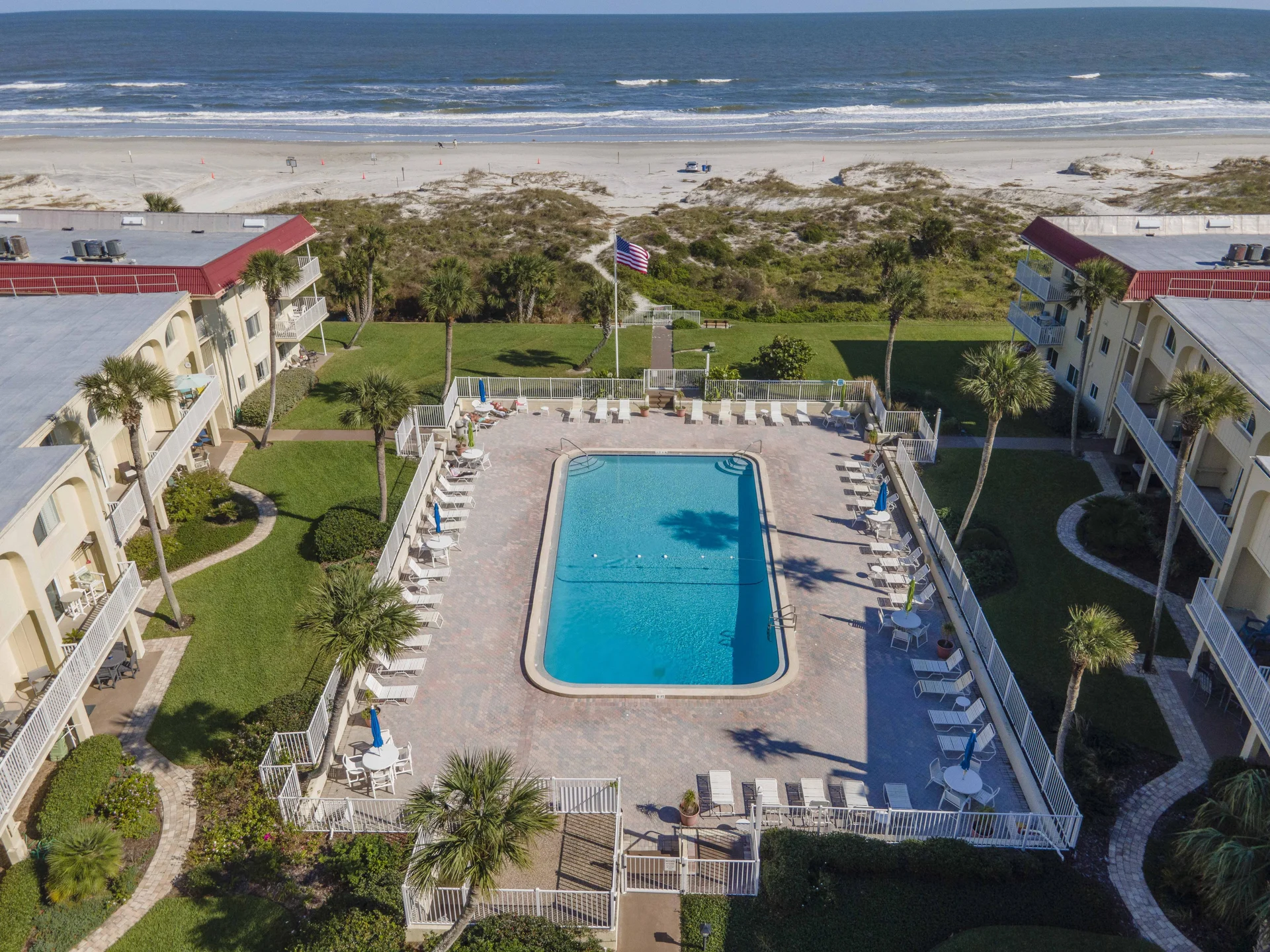 Rectangular pool with pale brick pavers is surrounded by white lounge chairs and fenced landscaping near the beach.