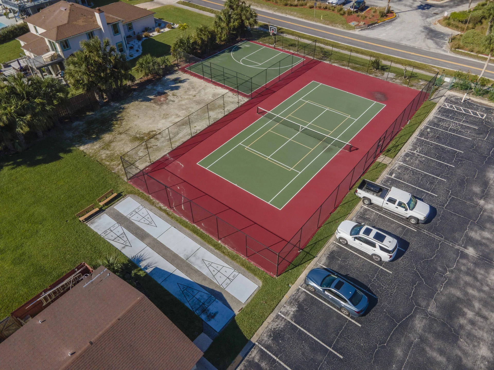 Exterior view of fenced tennis and basketball courts with red and green surfaces adjacent to shuffleboard courts and parking.