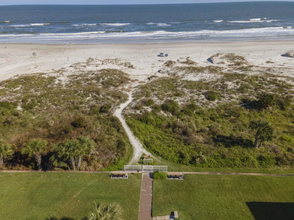 A paved pathway leads from green lawn past palm trees and dunes to a sandy beach with gentle ocean waves.