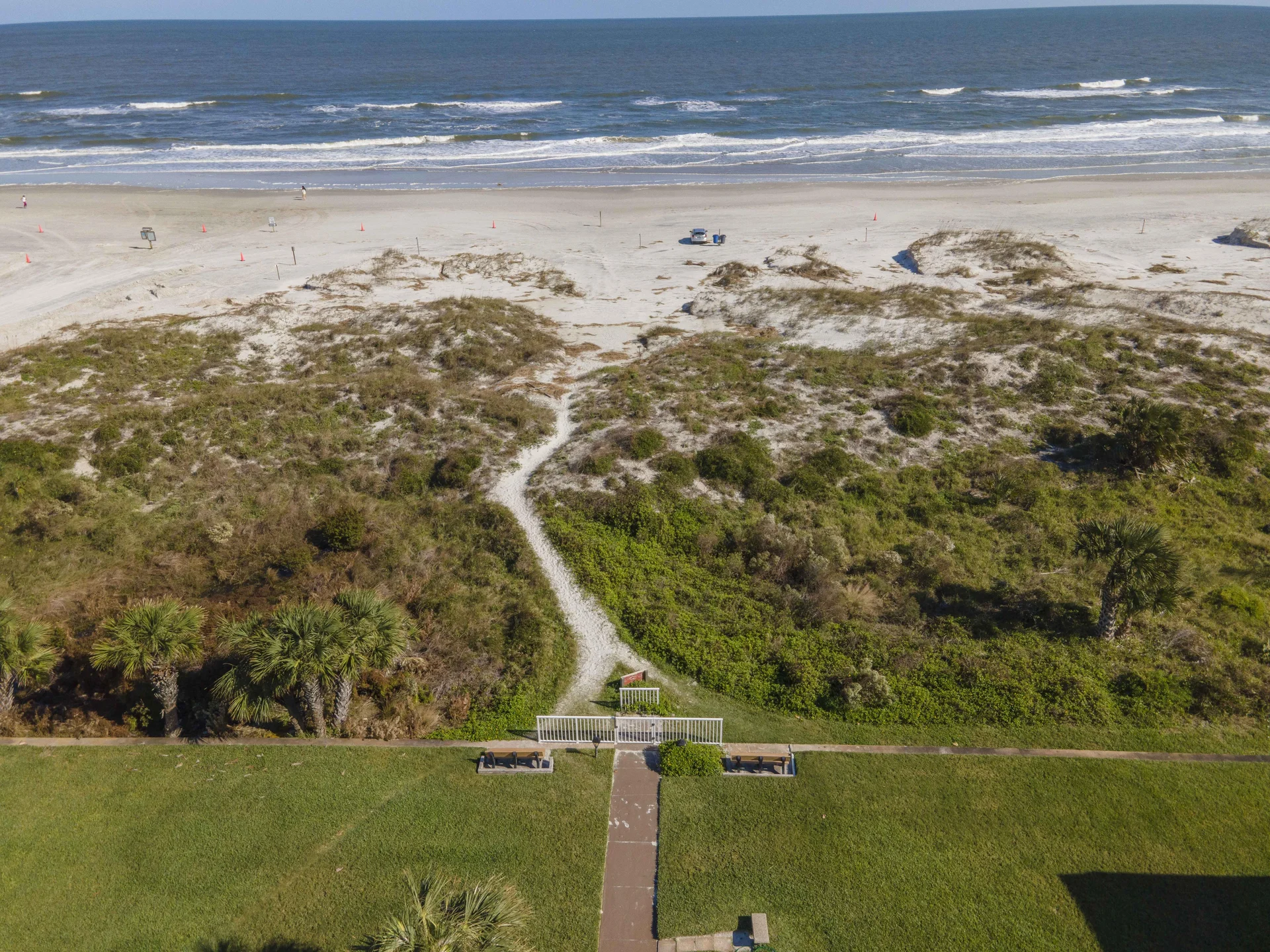 A paved pathway leads from green lawn past palm trees and dunes to a sandy beach with gentle ocean waves.