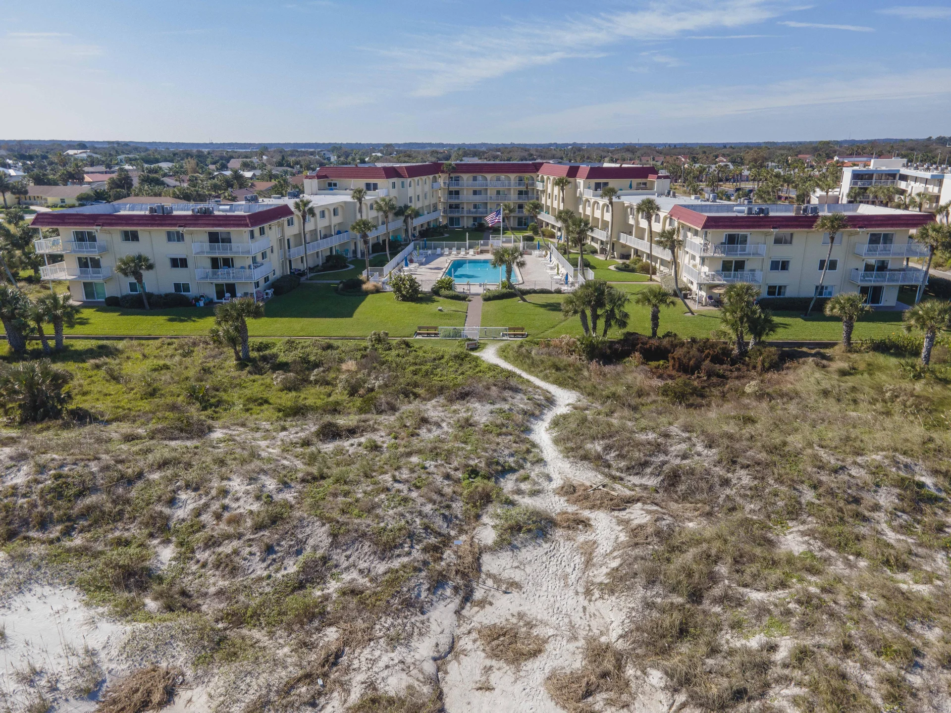 Exterior view of three-story beige condo buildings with red roofs surrounding a fenced pool, grassy lawn, and palm trees.