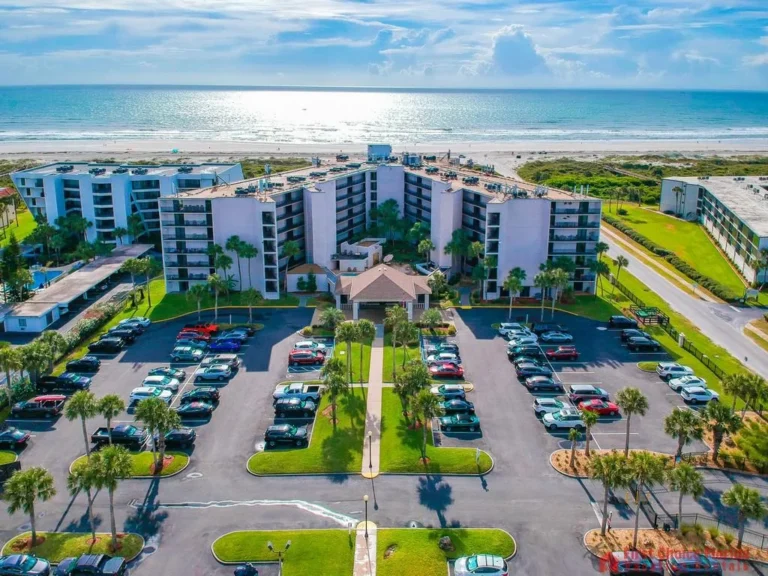 Aerial view of a large condo complex with multiple parking rows, palm trees, and the ocean beyond a sandy beach.