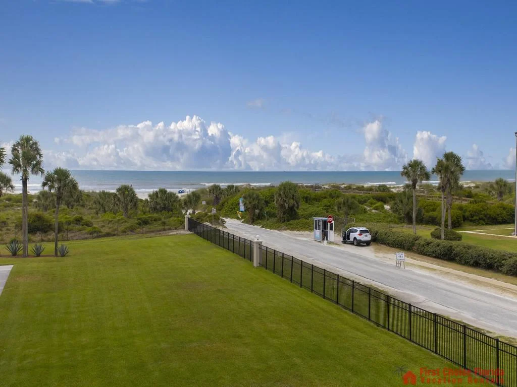 Oceanfront view over a manicured lawn and black metal fence, with palm trees and a sandy beach beyond.