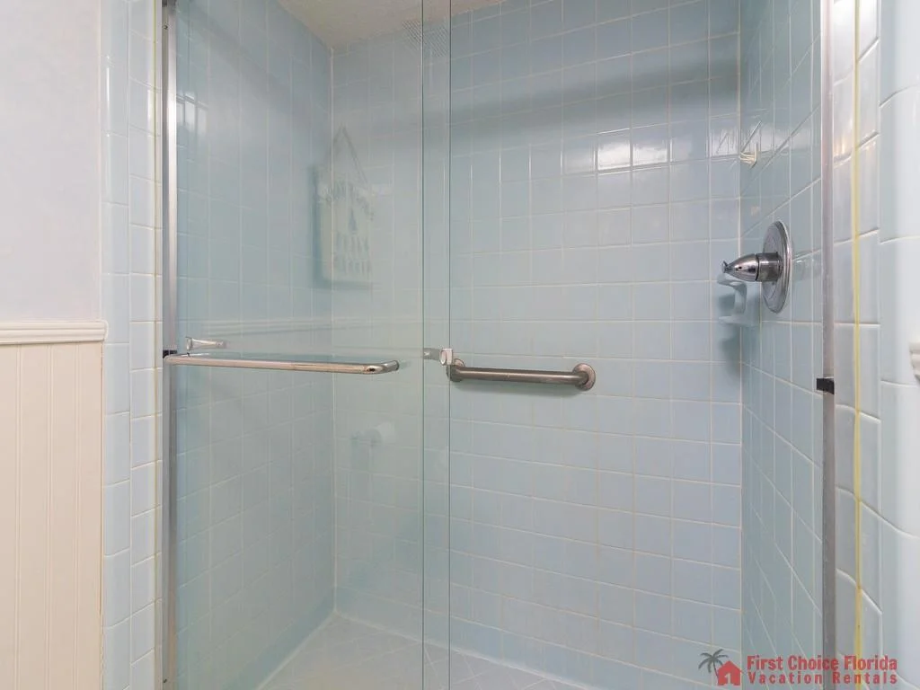 Glass-enclosed shower features light blue square tiles, a chrome grab bar, and a single-handle faucet.