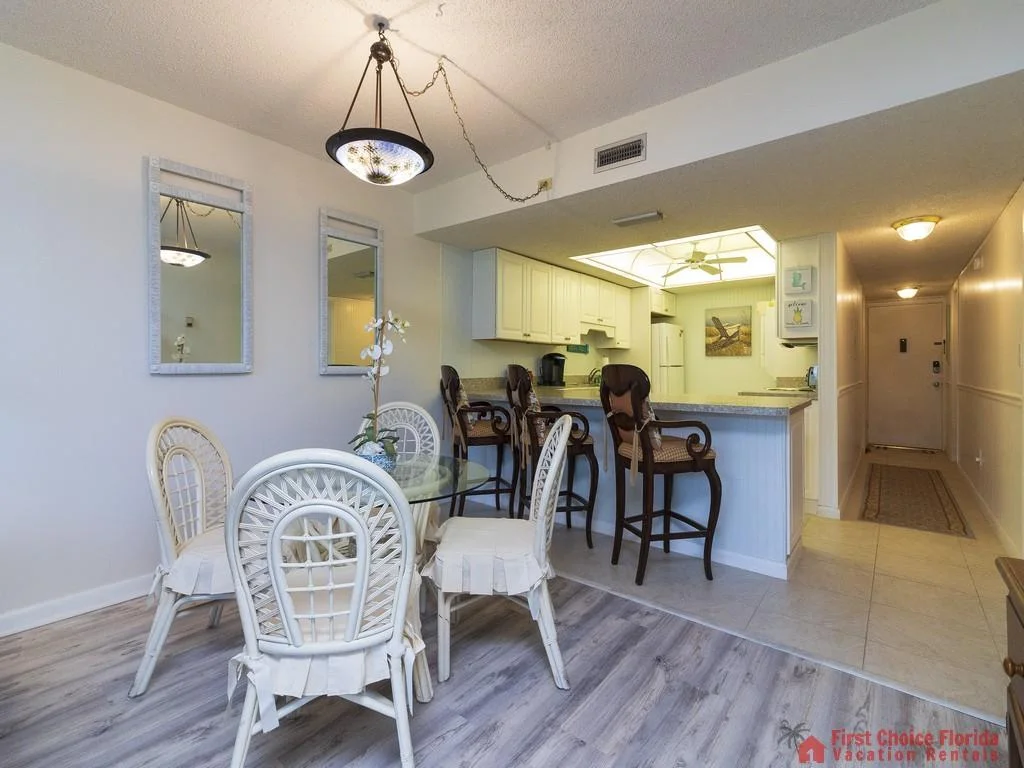 Glass-top dining table surrounded by white wicker chairs sits beside a breakfast bar with ornate wooden stools.